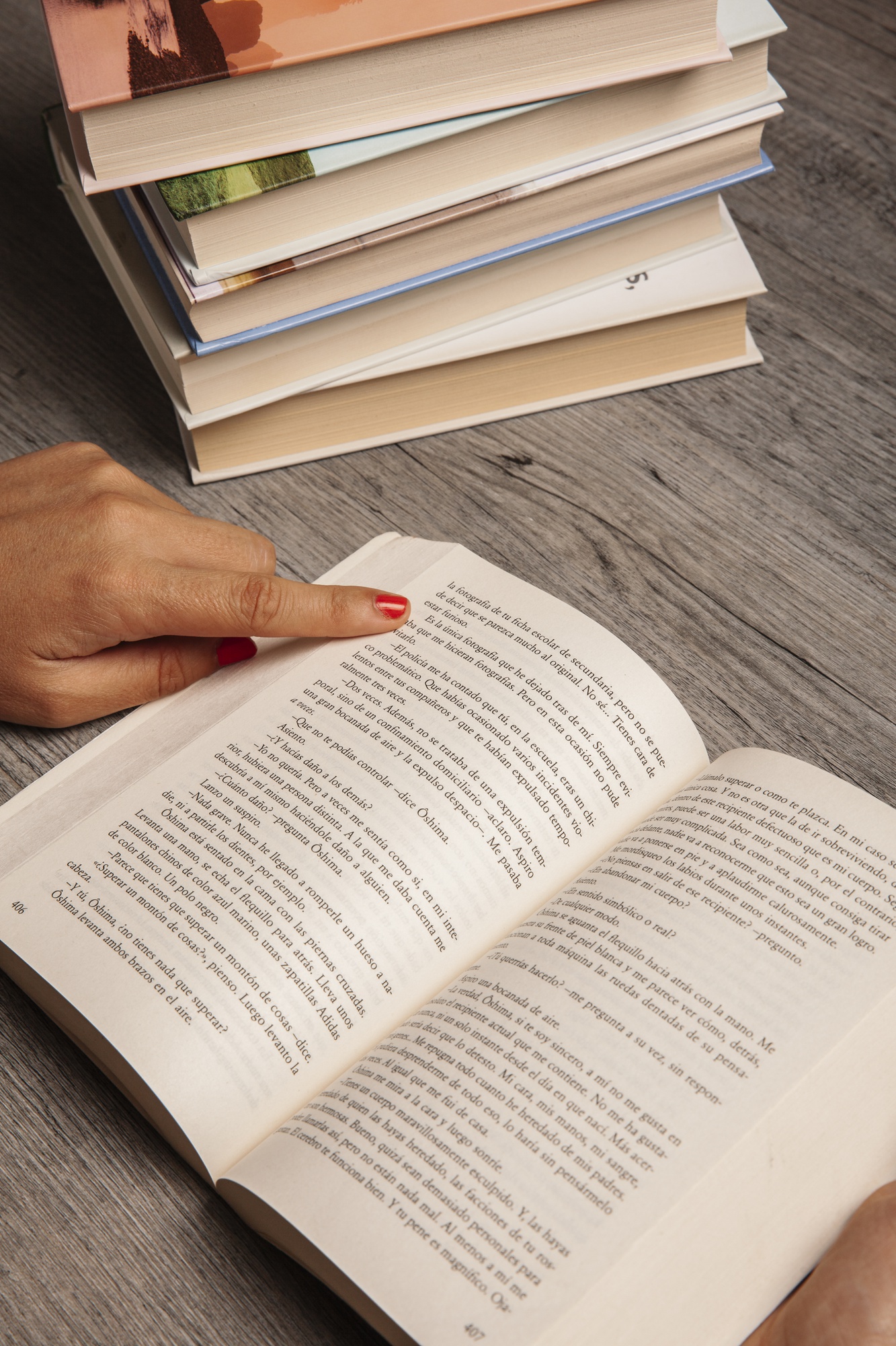 Mão apontando para o texto de um livro aberto, com uma pilha de livros ao fundo sobre uma mesa de madeira.